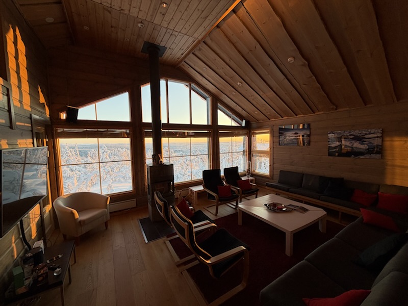 Salon du chalet, grandes baies vitrées sur la forêt enneigée au coucher du soleil